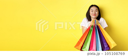 Happy asian female shopper smiling and holding colorful shopping bags, standing against yellow background 105108769