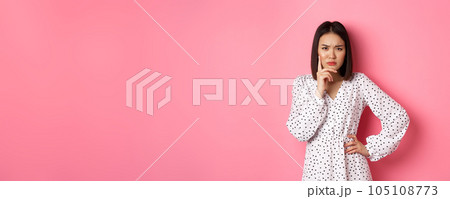 Concerned asian woman staring at camera suspicious, having doubts, thinking about something with serious face, standing against pink background Concerned asian woman staring at camera suspicious, having doubts, thinking about something with serious face, standing against pink background 105108773