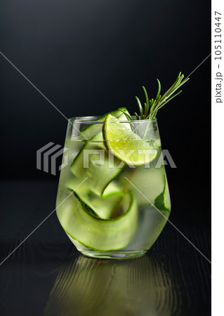 Gin tonic with ice, rosemary, lime, and cucumber in frosted glass on a black background. 105110447