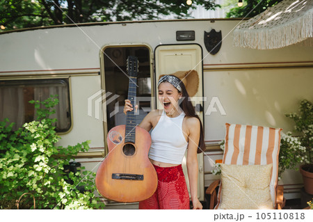 Happy hippie girl is having a good time with playing on guitar in camper trailer. Holiday, vacation, trip concept. 105110818