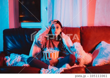 A gamer or a streamer girl at home in a dark room with a popcorn bucket playing with her dog and sits in front of a monitor or TV. A gamer or a streamer girl at home in a dark room with a popcorn bucket playing with her dog and sits in front of a monitor or TV. 105111061