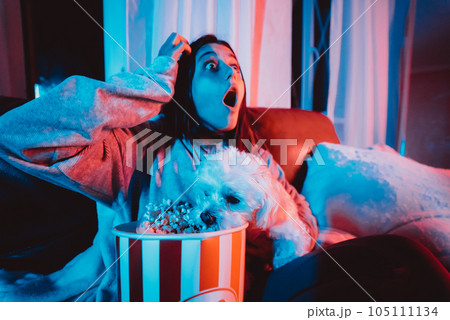 Close up portrait of Beautiful young girl at home in a dark room with a popcorn bucket playing with her dog and sits in front of a monitor or TV. 105111134