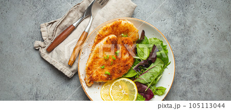 Crispy panko breaded fried chicken fillet with green salad and lemon on plate with cutlery on gray rustic concrete background table from above. Japanese style deep fried coated chicken breasts . Crispy panko breaded fried chicken fillet with green salad and lemon on plate with cutlery on gray rustic concrete background table from above. Japanese style deep fried coated chicken breasts . 105113044