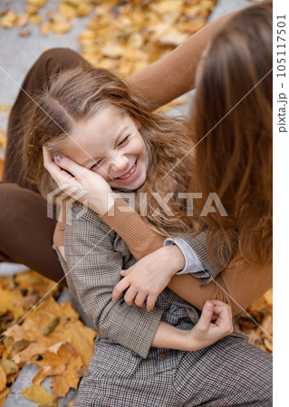 Young woman and little girl in autumn forest. Woman hugging her daughter. Girl wearing fashion clothes and lying on mother's lap. 105117501