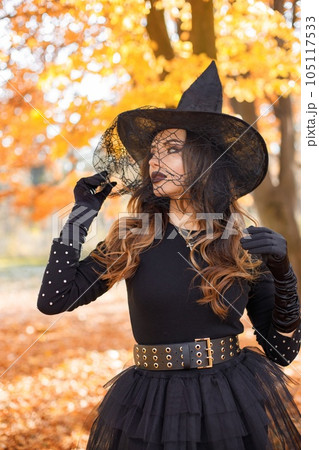 Brunette woman in witch costume standing in autumn forest on Halloween day. Woman wearing black clothes and cone hat. Woman posing for a photo. 105117533