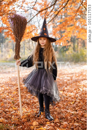 Cute happy little girl dressed in witch costume standing with over autumn forest background. Girl wearing dress and cone hat and holding a broom. Girl dressed for Halloween. Cute happy little girl dressed in witch costume standing with over autumn forest background. Girl wearing dress and cone hat and holding a broom. Girl dressed for Halloween. 105117553