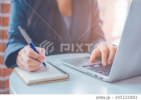 Woman hand is writing on a notepad with a pen and have a laptop computer on the desk in the office. 105122001