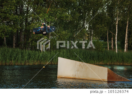 Beautiful view of young man rider holding rope and making extreme jump on wakeboard. Wakeboarding and water sports activity. Low angle shot of man wakeboarding on a lake. Man water skiing at sunset. 105126374