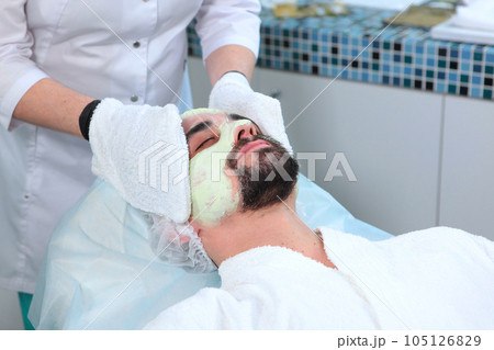 Men's cosmetology. The cosmetologist removes a nourishing mask from the man's face to moisturize the skin. Portrait of a man with a clay face mask. The procedure in beauty salon. Men's cosmetology. The cosmetologist removes a nourishing mask from the man's face to moisturize the skin. Portrait of a man with a clay face mask. The procedure in beauty salon. 105126829