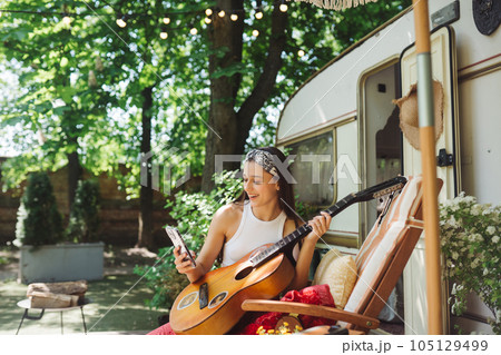 Happy hippie girl is having a good time with playing on guitar with smartphone in her hand in camper trailer. Holiday, vacation, trip concept. Happy hippie girl is having a good time with playing on guitar with smartphone in her hand in camper trailer. Holiday, vacation, trip concept. 105129499