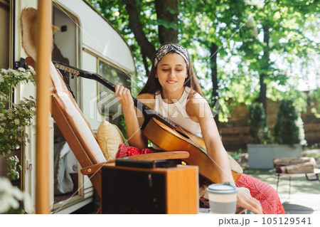Happy hippie girl is having a good time with playing on guitar with smartphone in her hand in camper trailer. Holiday, vacation, trip concept. 105129541