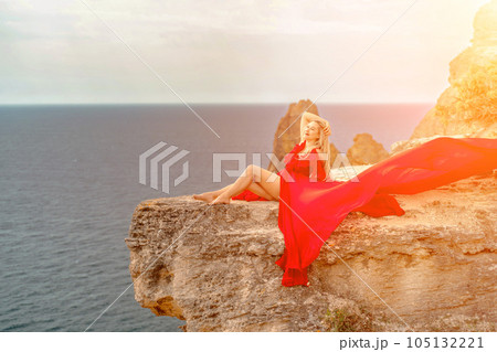 woman red silk dress sits by the ocean with mountains in the background, her dress swaying in the wind. 105132221