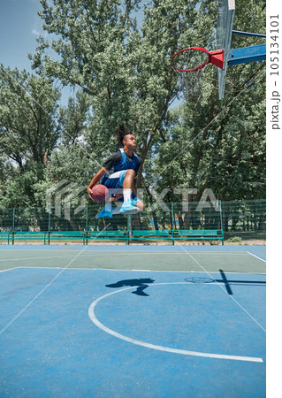 Slam dunk. Full-length image of young man in motion, playing basketball outdoors on sportsground on warm day. 105134101