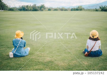 Two women sitting on the grass field, looking to beautiful view and keep space for avoid spreading of virus disease in covid-19 pandemic. 105137242
