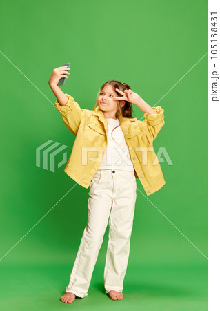 Full-length shot of little girl dressed stylishly taking selfie on cell phone and showing v-sign isolated on green background. Full-length shot of little girl dressed stylishly taking selfie on cell phone and showing v-sign isolated on green background. 105138431