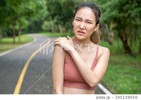 Young atractive Asian woman in fitness clothes putting her left hand over her pain right shoulder while standing at running track of a local park 105139050