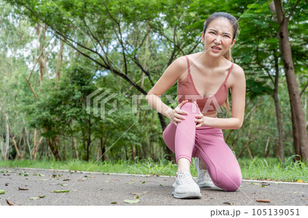 Young atractive Asian woman with knee pain putting her hands over her troubled right knee while sitting down at running track of a local park 105139051