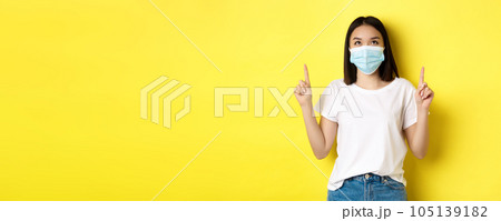Covid-19, pandemic and social distancing concept. Young asian woman in white t-shirt and medical mask from coronavirus, looking and pointing fingers up, showing special offer 105139182