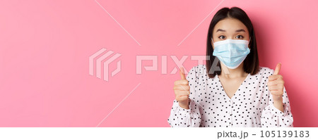 Covid-19, pandemic and lifestyle concept. Supportive asian female in face mask showing thumbs-up, approve and praise good work, standing over pink background Covid-19, pandemic and lifestyle concept. Supportive asian female in face mask showing thumbs-up, approve and praise good work, standing over pink background 105139183