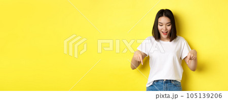 Beauty and fashion concept. Beautiful asian woman in white t-shirt pointing fingers down, demonstrate logo standing over yellow background Beauty and fashion concept. Beautiful asian woman in white t-shirt pointing fingers down, demonstrate logo standing over yellow background 105139206
