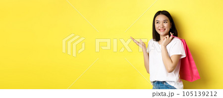 Cheerful asian female shopper looking amused, holding shopping bag and pointing hand left at store banner, standing over yellow background 105139212