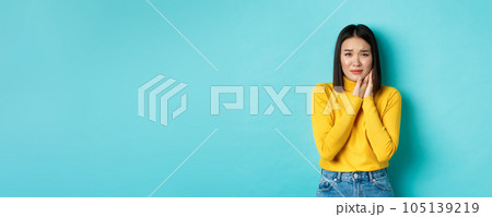 Young asian woman need dentist, touching cheek and frowning from painful toothache, standing over blue background. Health care concept 105139219