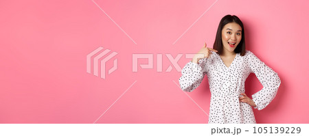 Beautiful asian girl in dress looking happy, pointing finger at herself, standing on romantic pink background Beautiful asian girl in dress looking happy, pointing finger at herself, standing on romantic pink background 105139229