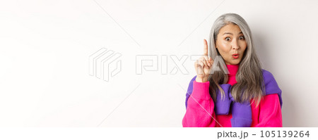 Close up of elderly asian woman in stylish hipster clothes, raising finger and saying idea, have suggestion, standing over white background 105139264