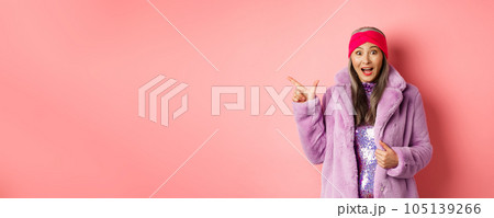 Fashion and shopping concept. Fashionable senior asian woman in winter coat and headaband pointing finger left at copy space, staring amazed at camera, checking out banner Fashion and shopping concept. Fashionable senior asian woman in winter coat and headaband pointing finger left at copy space, staring amazed at camera, checking out banner 105139266