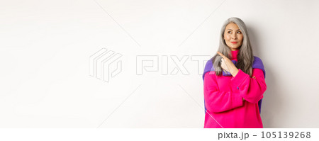 Stylish modern asian elderly woman pointing at upper left corner, looking at logo with pleased smile, standing in pink sweater over white background Stylish modern asian elderly woman pointing at upper left corner, looking at logo with pleased smile, standing in pink sweater over white background 105139268