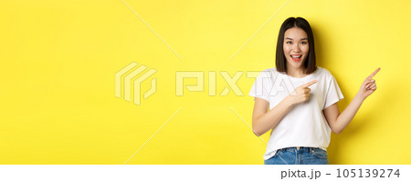 Beauty and fashion concept. Beautiful asian woman in white t-shirt pointing fingers right, demonstrate logo standing over yellow background Beauty and fashion concept. Beautiful asian woman in white t-shirt pointing fingers right, demonstrate logo standing over yellow background 105139274