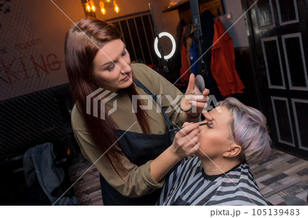 A cute attractive adult girl, an experienced salon worker, makes make-up with shadows, palette, paints eyelids with a brush, eyes to the client, services 105139483