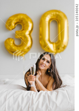 Happy young woman meets her thirtieth birthday in bed, holding a glass of champagne in her hands and smiling 105140042