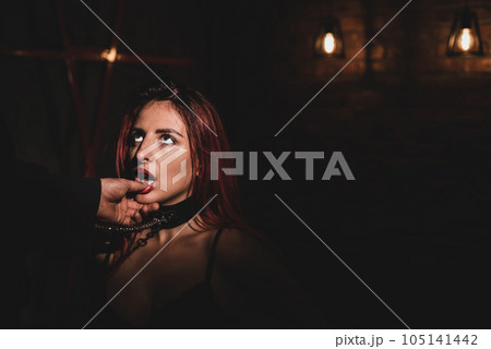 BDSM. Portrait of a red-haired woman in shackles kneeling and licking the fingers of a man. 105141442