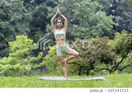 Young beautiful woman in the park on a sports mat doing fitness and doing active sports exercises, Hindu woman doing yoga in the park in the morning among the trees, stretching and meditating. 105141515