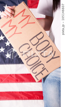 Young woman protester holds cardboard with My Body My Choice sign against USA flag on background. Girl protesting against anti-abortion laws. Feminist power. Equal opportunity Womens rights reedom. Young woman protester holds cardboard with My Body My Choice sign against USA flag on background. Girl protesting against anti-abortion laws. Feminist power. Equal opportunity Womens rights reedom. 105146607