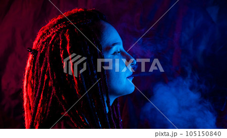 Profile of caucasian girl with dreadlocks smokes a vape in red blue light.  105150840