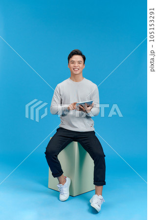 Full length view of young man in jeans and gray t-shirt sitting cube on blue background Full length view of young man in jeans and gray t-shirt sitting cube on blue background 105153291