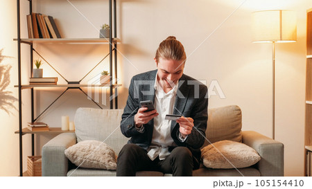 Mobile cashback. Bank app. Successful electronic transaction. Satisfied excited business man using credit card phone for virtual payment on couch at home interior with free space. 105154410