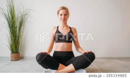 Morning yoga. Fitness leisure. Pilates workout. Smiling woman sitting cross-legged in lotus position on floor at light home interior with free space. 105154903