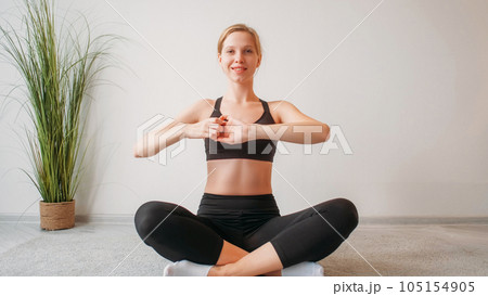 Indoor fitness. Home training. Yoga exercise. Smiling athletic woman enjoying stretching arms sitting cross-legged on floor at light studio interior with copy space. 105154905