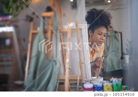 Charming female artist sitting in front of an easel and dipping brush into color palette 105154910