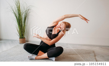 Home pilates. Yoga exercise. Indoor fitness training. Relaxed athletic woman stretching back arms muscles sitting cross-legged on floor at light studio interior with free space. 105154911