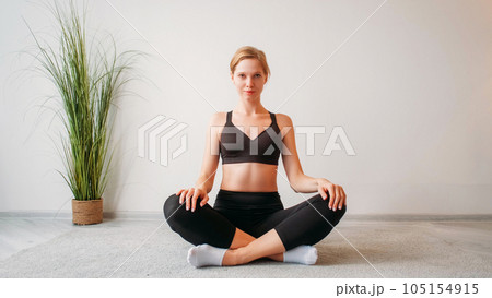 Indoor yoga. Home training. Health wellbeing. Confident relaxed woman sitting cross-legged in lotus position on floor at light studio interior with empty space. 105154915