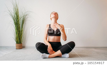 Morning exercise. Home fitness. Indoor training. Relaxed inspired athletic girl stretching neck muscles sitting cross-legged on floor at light studio interior copy space background. 105154917