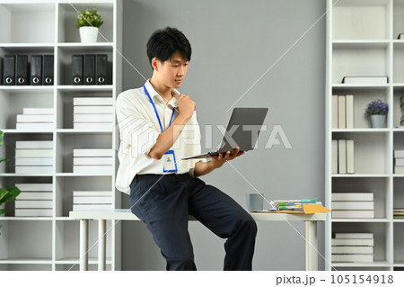 Focused Asian male manager sitting on desk in modern workplace and reading online information on laptop Focused Asian male manager sitting on desk in modern workplace and reading online information on laptop 105154918