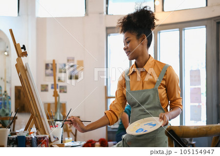 Creative young woman with Afro haired wearing apron painting picture with brush on canvas in art studio Creative young woman with Afro haired wearing apron painting picture with brush on canvas in art studio 105154947