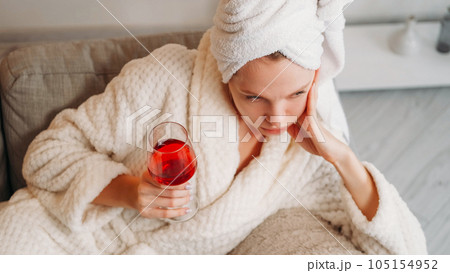 Lonely drink. Home depression. Boring weekend. Upset sad pensive woman in bathrobe with glass of alcohol red wine alone on couch after bath. Lonely drink. Home depression. Boring weekend. Upset sad pensive woman in bathrobe with glass of alcohol red wine alone on couch after bath. 105154952