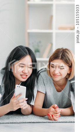 Device leisure. Technology youth. Two friends girls enjoying smartphone gadget internet social media content fun resting in light interior on floor at home. 105155033