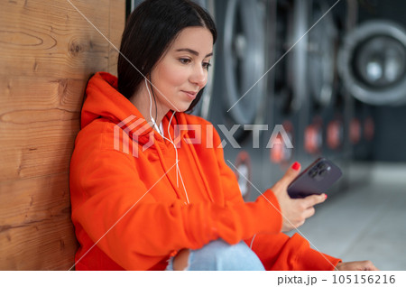 Young woman listening music with smartphone and earphones waiting in laundry room. Young woman listening music with smartphone and earphones waiting in laundry room. 105156216